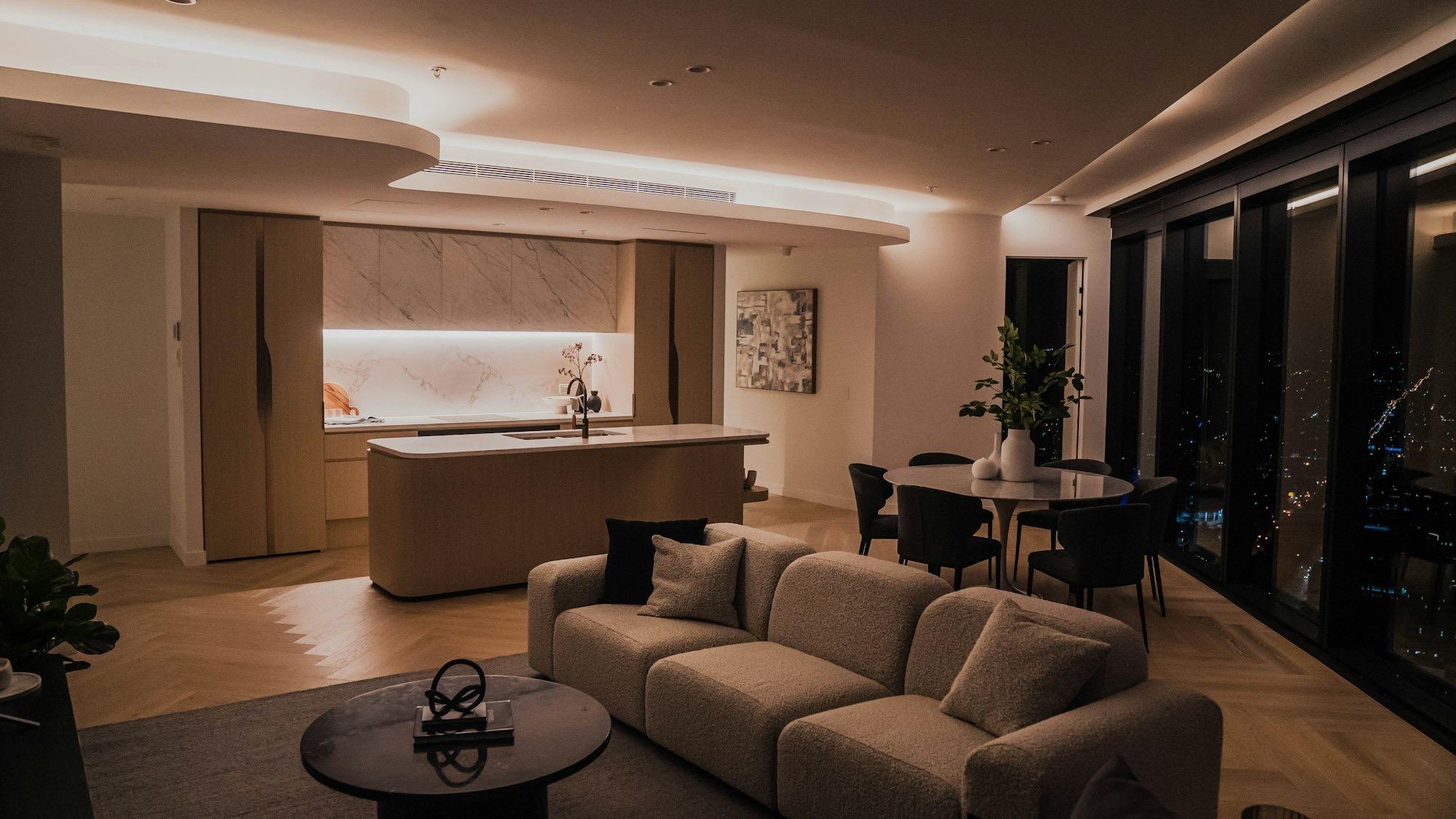 A high-rise apartment at night with a boucle sofa, marble kitchen island and floor-to-ceiling windows over city lights.