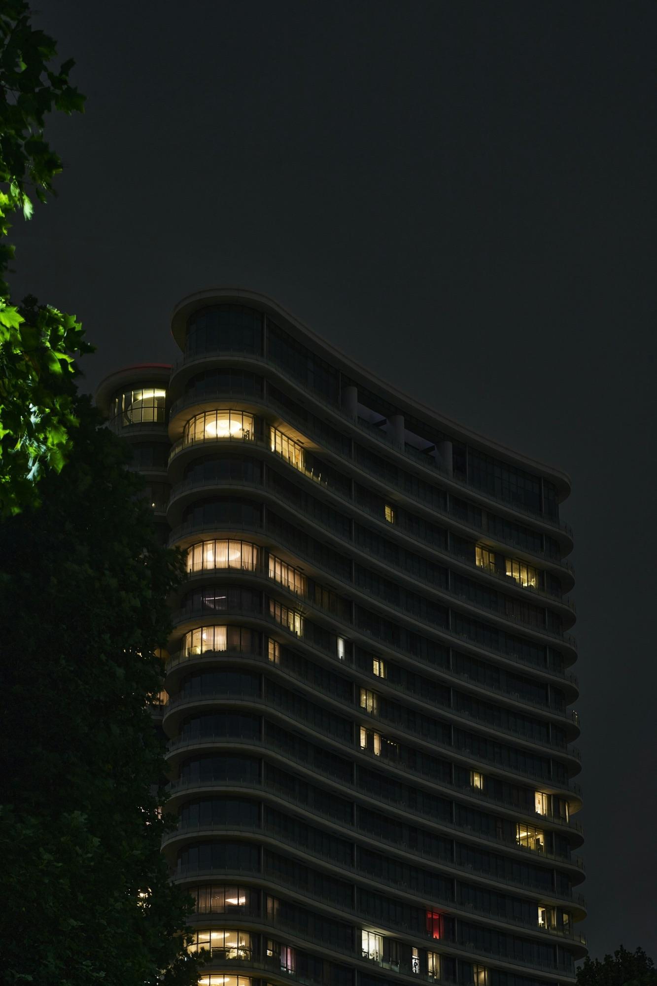 A curved modern residential tower at night, warm light glowing from a few windows against a dark sky.