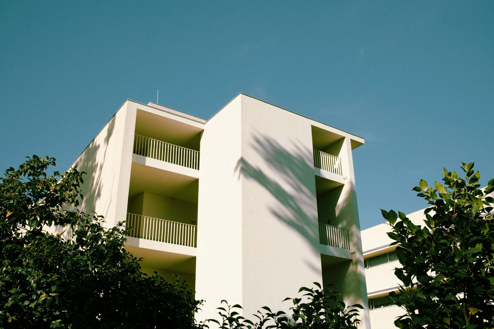 A white mid-rise apartment building with balconies, framed by leafy green trees under a clear blue sky.