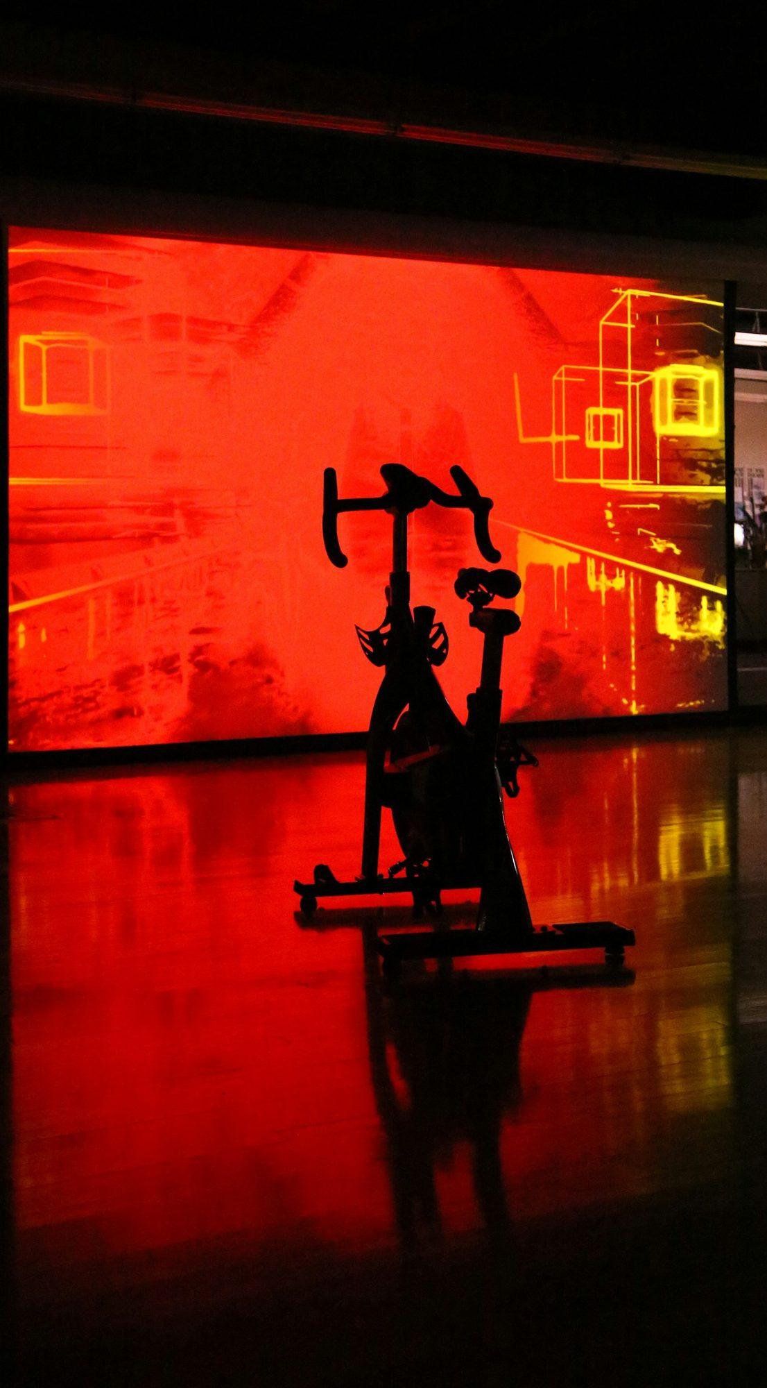 An indoor cycling bike silhouetted against a glowing screen in a dark studio.